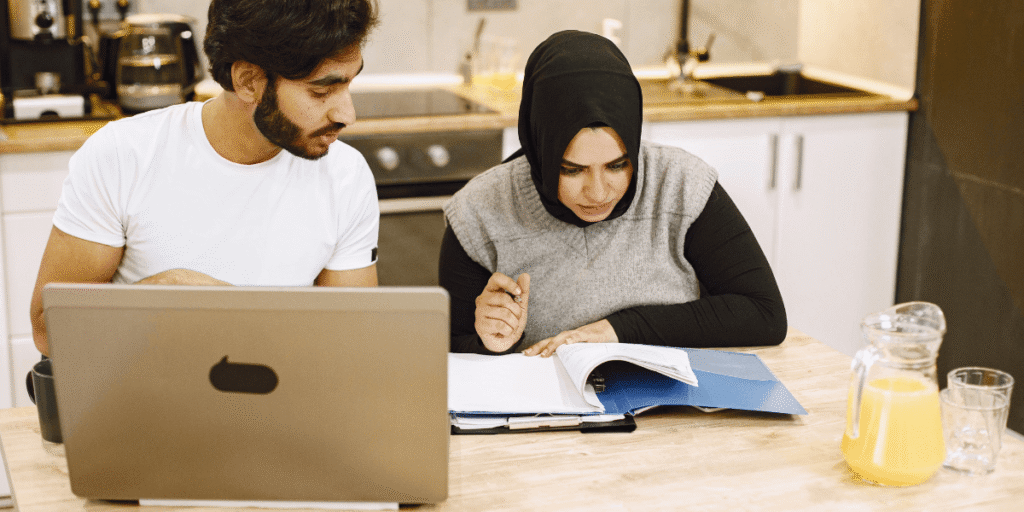 A man and woman in hijab sit at a table, focused on a laptop, engaged in a discussion or work.